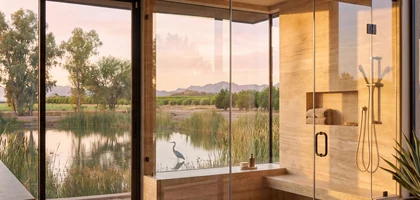 Frameless glass shower with a window view over a reed-lined pond where a heron stands, with mountains beyond