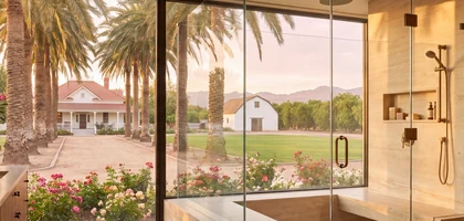 Frameless glass shower with a window view of palm trees, rose beds and a white ranch house set on a green lawn