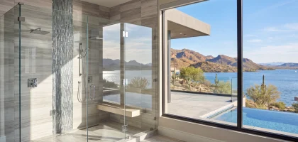 Frameless glass shower in gray stone tile, with a window overlooking a desert lake, an infinity pool and mountains