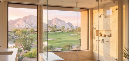 Frameless glass shower with a window overlooking a golf course fairway, palm trees and mountains at sunset