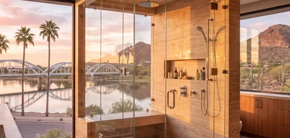 Frameless glass shower beside a window overlooking palm trees, a lake and an arched bridge at sunset